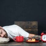Exhausted man asleep on a table with a coffee cup and alarm clock.